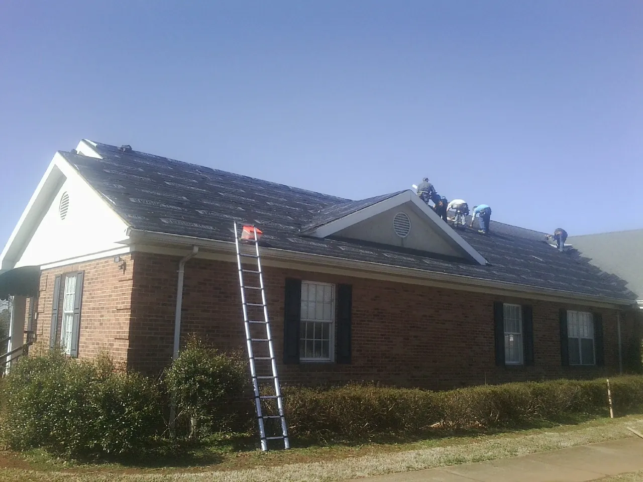 Crew tearing shingles off a brick ranch roof with a ladder set — Metal Roof Repair work by our crew in Manchester Center